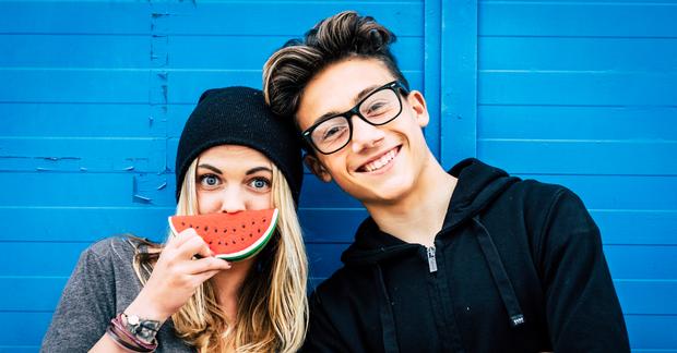 teen boy with glasses teen girl with watermelon slice in front of her mouth with blue painted wall